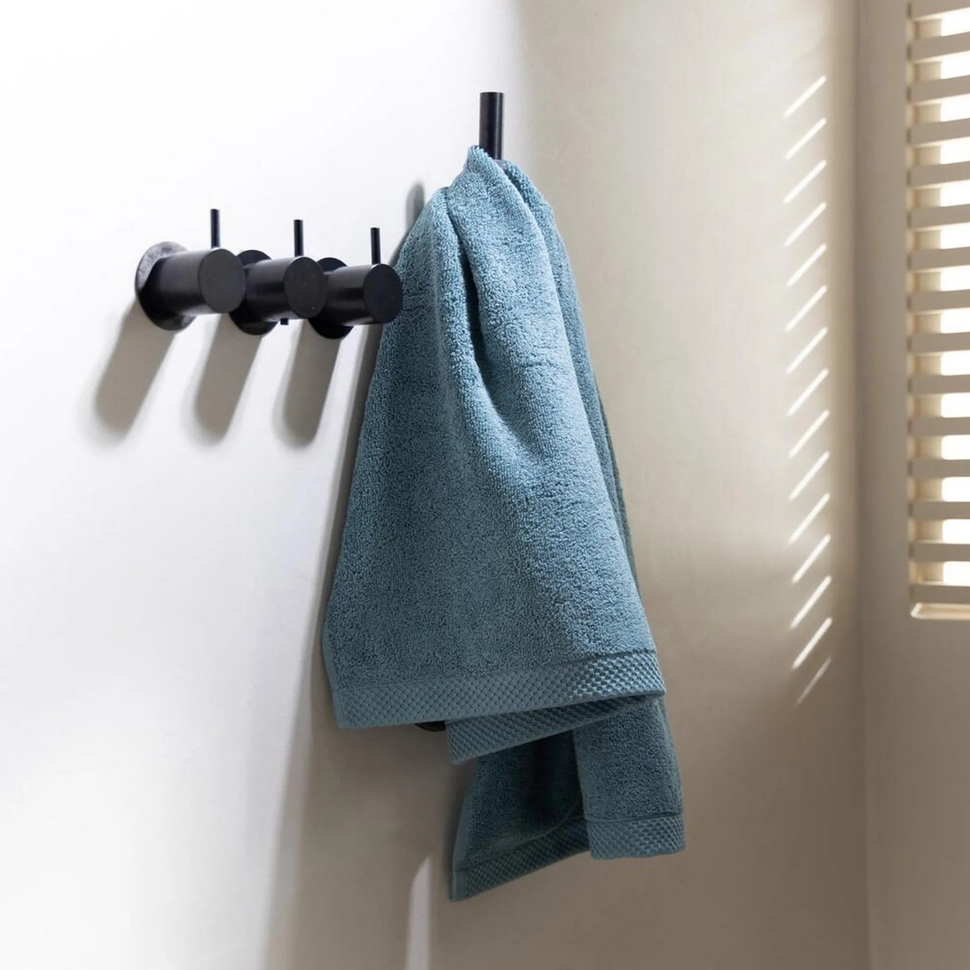 Gevouwen Florence-handdoek in mineraalblauw, hangend aan een van de vier haakjes in een badkamer