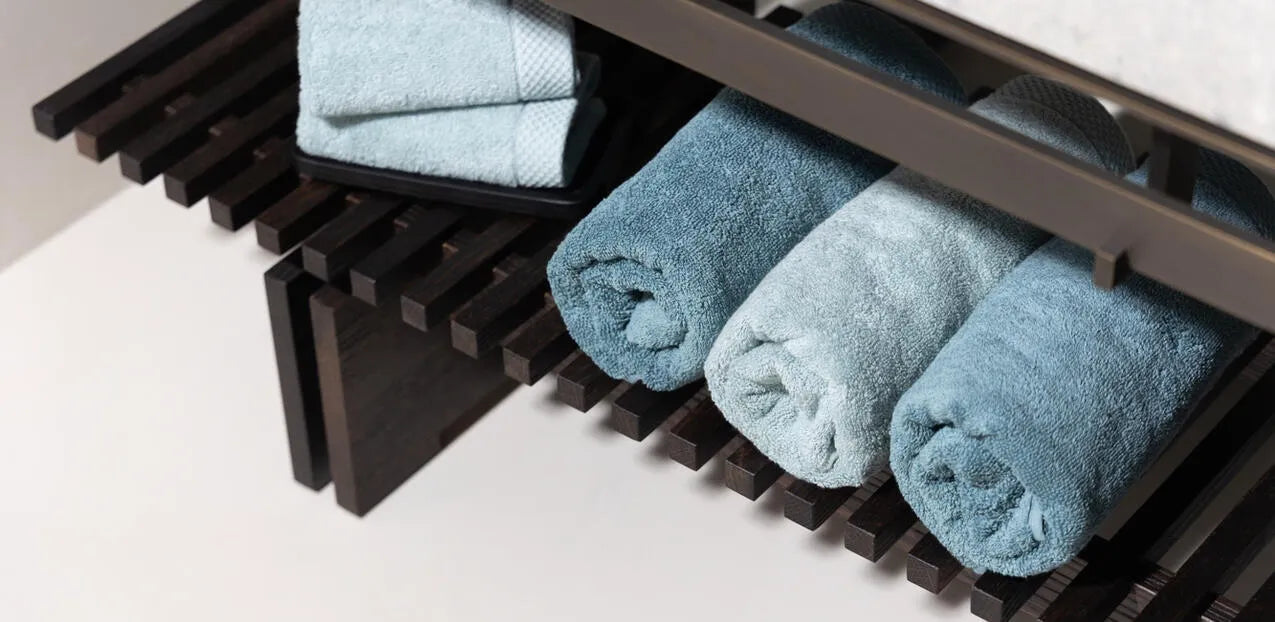 Drie opgerolde Florence-handdoeken in mineraalblauw, liggend naast elkaar in een open houten rek in de badkamer